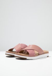 Pink slide sandals with a cross strap design, made of smooth and suede materials. Features a white sole with textured grip and a gold accent.