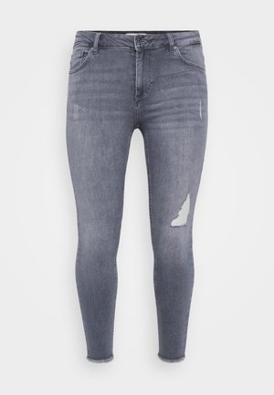 Jeans skinny en denim gris clair avec un léger délavage et une grande patch usé sur la jambe gauche, présentant un ourlet effiloché aux chevilles.