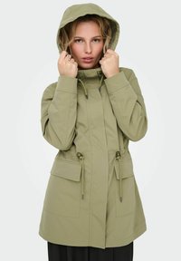 Veste imperméable vert olive avec capuche, dotée de cordons ajustables, de deux poches frontales et d'une coupe décontractée.