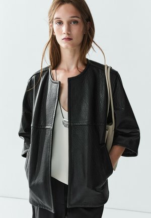 Junge Frau trägt eine schwarze Lederjacke mit viertellangen Ärmeln und ein weißes Top, und trägt eine beige Tasche auf der Schulter.