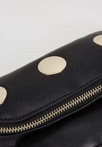 Detailní záběr na černou koženou psaníčko se zlatým zipem a třemi velkými kulatými zlatými nýty na klopě.