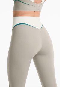 Close-up van een persoon die hoogpolige beige leggings draagt met een witte en teal tailleband en een bijpassende sportbeha.
