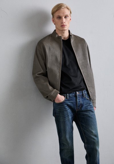 Pilkas tekstūruotas švarkas su pakeliamu apykakle, nešiojamas ant juodos marškinėlių, derinamas su mėlynomis džinsinėmis kelnėmis. Minimalistinis dizainas be matomų logotipų.