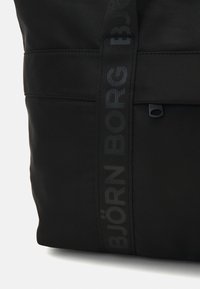 Saco tote preto com um tecido texturizado, apresentando uma alça tecida com a inscrição 'BJÖRN BORG' em uma impressão discreta e um pequeno bolso com fecho éclair.