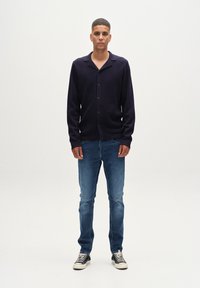 Mornarsko rebrasto cardigan s gumbom, oblečen nad modrimi kavbojkami in črnimi supergami. Model stoji pred enobarvno belo ozadje.