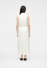 Femme aux longs cheveux foncés portant une robe midi blanche sans manches et des chaussures à talons noirs, vue de dos sur un fond blanc uni.