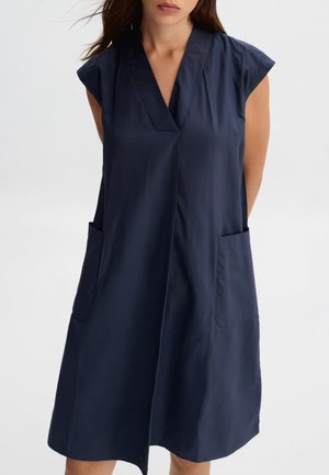 Donna con vestito blu navy senza maniche, lunghezza al ginocchio, con scollatura a V profonda e due tasche frontali, in piedi con le mani dietro la schiena.