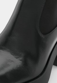Black ankle boot made of shiny leather with elastic side panels and a chunky heel. Smooth surface, rounded toe, and minimal stitching detail.