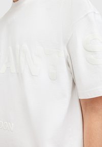 White cotton t-shirt with a crew neck, featuring raised, textured lettering that reads "SAINTS LONDON" on the front and sleeve.