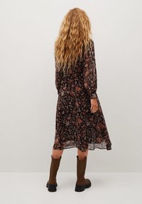 Blumenkleid mit schwarzem Hintergrund und bunten Blumen, langen Ärmeln und knielangem Saum. Kombiniert mit braunen Stiefeln bis zum Knie.