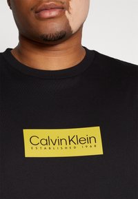 Černé tričko s výrazným žlutým obdélníkovým logem s textem "Calvin Klein Established 1968" v černé barvě. Bavlněný materiál s žebrovaným výstřihem.