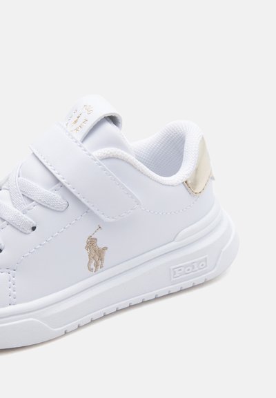 Zapatilla blanca Polo Ralph Lauren con bordado de logo en dorado, cierre de velcro y collar acolchado, mostrada desde un ángulo lateral y trasero.