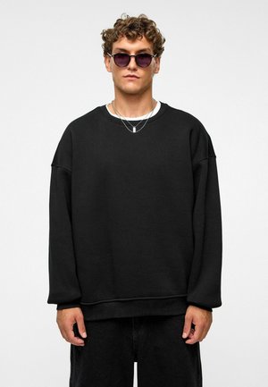 Zwarte oversized sweatshirt met een ronde hals, gemaakt van zachte stof. Model draagt zilveren kettingen en ronde zonnebril. Eenvoudig, casual ontwerp.