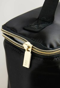 Borsa in nylon nera con una cerniera dorata. Presenta una maniglia robusta e una consistenza liscia. Vista ravvicinata del tirante della cerniera con la lettera incisa 'B'.