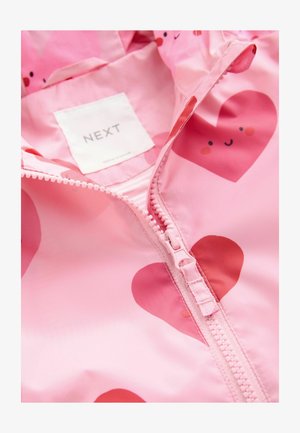 Rožinė neperšlampama striukė su širdelių raštu, priekyje su užtrauktuku ir etiketė su užrašu „NEXT“. Lygi tekstūra su žaismingais akcentais.