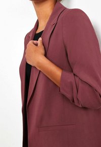 Maroon blazer dengan potongan yang disesuaikan, dilengkapi dengan kerah bergerigi, saku dada tunggal, dan lengan yang digulung. Tekstur kain yang halus.