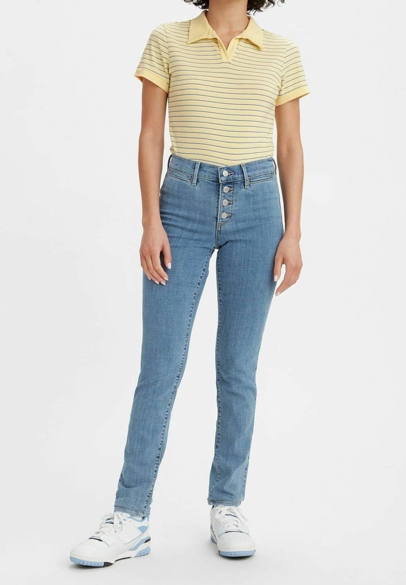 Femme portant un polo rayé jaune, un jean taille haute bleu avec des boutons, et des baskets blanches, debout devant un fond uni.