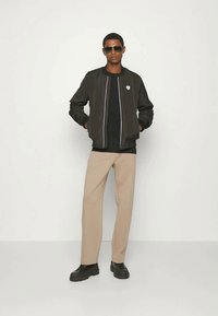 Homem a usar uma jaqueta bomber preta, camisa preta, calças bege, sapatos pretos e óculos de sol, em pé contra um fundo branco.