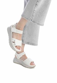 Sandalias de cuña blancas con correas perforadas, cierres ajustables en el tobillo y en la parte delantera del pie, y suela acolchada, mostradas con pantalones anchos grises.