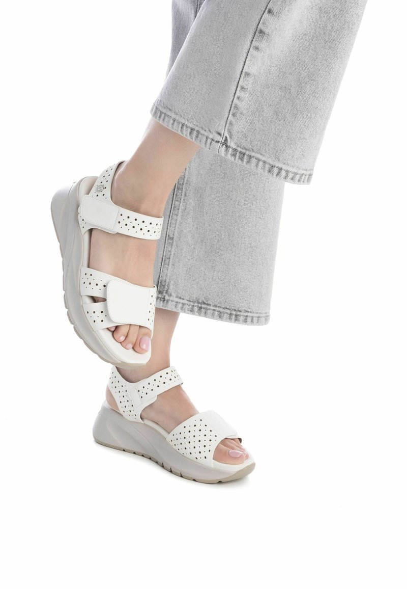 Sandalias de cuña blancas con correas perforadas, cierres ajustables en el tobillo y en la parte delantera del pie, y suela acolchada, mostradas con pantalones anchos grises.