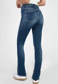 Uitlopende blauwe jeans van denim met een middelhoge taille, vijf zakken en zichtbare stikdetails, gecombineerd met witte sneakers.