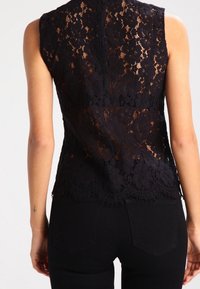 Svart spetslinne utan ärmar med blommönster, vågig fåll och en texturerad design, tillsammans med mörka jeans.