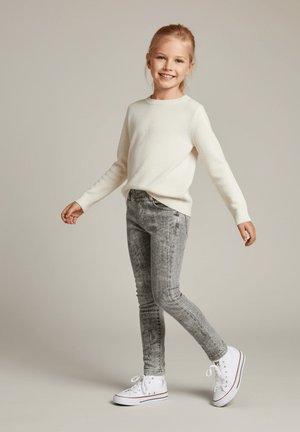 Uśmiechnięta młoda dziewczyna o blond włosach, ubrana w biały sweter, szare jeansy z efektem sprania i białe trampki, stojąca na neutralnym tle.