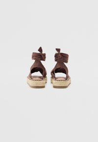 Espadrile din material textil maro, cu bretele legate, toc deschis și talpă din iută țesută, având un design plat și o textură netedă.