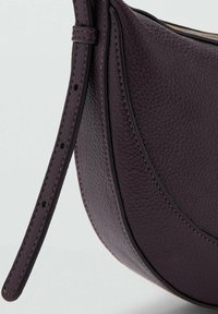 Bolso de cuero morado con una superficie texturizada, forma curva y costuras en contraste. Cuenta con una correa ajustable y bordes suaves.