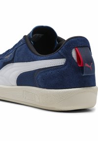 Marineblå ruskinds sneaker med hvide læderdetaljer, struktureret gummisål, sort foring og rødt Puma-logo på hælen.