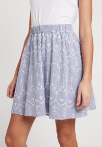Striped A-line skirt featuring a smocked waistband and intricate white embroidery of floral patterns. Made from lightweight fabric in blue and white.