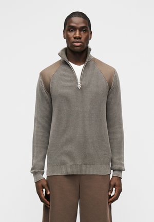 Mężczyzna ubrany w taupe sweter z prążkowanego dzianego materiału z ćwiartkowym suwakiem i kontrastującymi łatami na ramionach, w komplecie z dopasowanymi brązowymi spodniami, stoi nieruchomo.