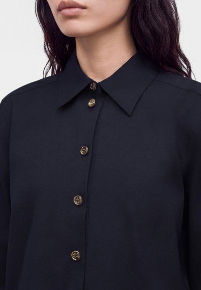 Jeune femme portant une chemise noire à col avec six boutons décoratifs dorés sur le devant, montrée des épaules au menton.