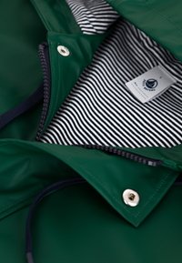 Veste imperméable verte avec doublure en tissu à rayures, fermeture éclair et accents en métal argenté. Équipée d'une capuche à cordon et d'une étiquette intérieure.