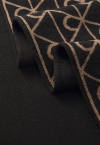 Svart stoff med beige geometrisk mønster brettet for å vise tekstur og strikkedetaljer.