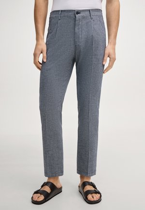 Homme portant un pantalon gris sur mesure et des sandales noires, debout devant un fond uni, les mains détendues le long du corps.