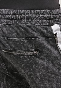 Černé joggingové kalhoty s popraskanou texturou, vybavené kapsou na zip a elastickým pasem. Doplňky zahrnují kontrastní boční panel.