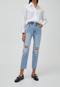 Witte lange mouwen geknoopte blouse in combinatie met lichtblauwe ripped jeans en zwarte instapschoenen met een gouden kettingaccent.