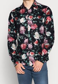 Man med svart långärmad skjorta med knäppning och blommönster i rött, rosa och vitt samt blå byxor, mot en enfärgad bakgrund.