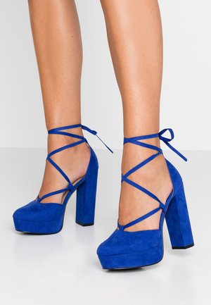 Niebieskie zamszowe szpilki na platformie z grubym słupkowym obcasem, otwartymi bokami i krzyżującymi się paskami na kostkach. Gładka tekstura o matowym wykończeniu.
