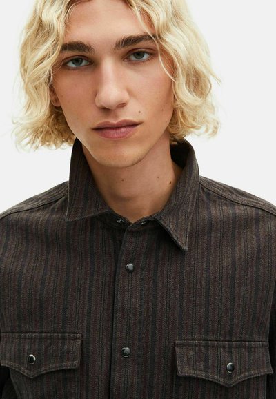 Νεαρό άτομο με κυματιστά ξανθά μαλλιά, φόρεμα με σκούρα ριγέ πουκάμισο με δύο τσέπες στο στήθος, σε απλό φόντο.