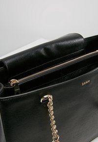 Sac à main en cuir noir à finition texturée, avec une poche zippée dorée et une bandoulière chaîne. La doublure intérieure est à motif.