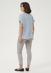 Top bleu à manches courtes avec un motif géométrique blanc, associé à un jean gris slim-fit et des chaussures plates noires. Tissu lisse ; design décontracté.
