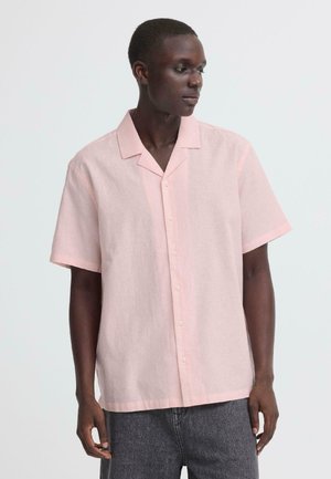 Ung mand iført en løs, lyserød kortærmet skjorte med knapper og mørkegrå bukser, kiggede mod højre.