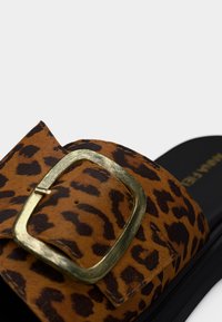 Κοντινό πλάνο του λουριού σανδάλιου με τύπωμα λεοπάρδαλης και μεγάλο χρυσό κουμπί πάνω σε μαύρη σόλα, με φόντο λευκό.