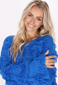 Königsblauer Pullover mit strukturierten Fransen an Ärmeln und Körper, der einen lockeren Schnitt und weite Bündchen aufweist. Das Material wirkt gestrickt und weich.