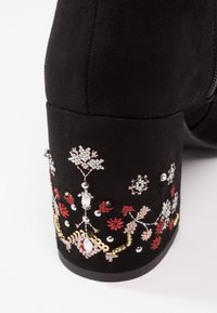 Bottine en daim noir à talon bloc haut, ornée d'une broderie florale complexe en rouge, or et argent sur le talon.