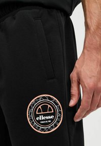 Pantalones de chándal negros con cintura elástica y bolsillos laterales, que presentan un logotipo circular en blanco y melocotón en el muslo.