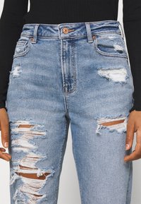 Džínové kalhoty světle modré barvy s odřenými detaily, obsahující potrhané části a roztrhané okraje. Klasický design s pěti kapsami a vysokým pasem.