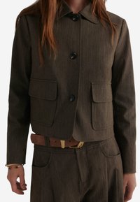 Braune maßgeschneiderte Jacke mit Kragen, ausgestattet mit zwei vorderen Taschen und Knopfverschluss, aus strukturiertem Material gefertigt.